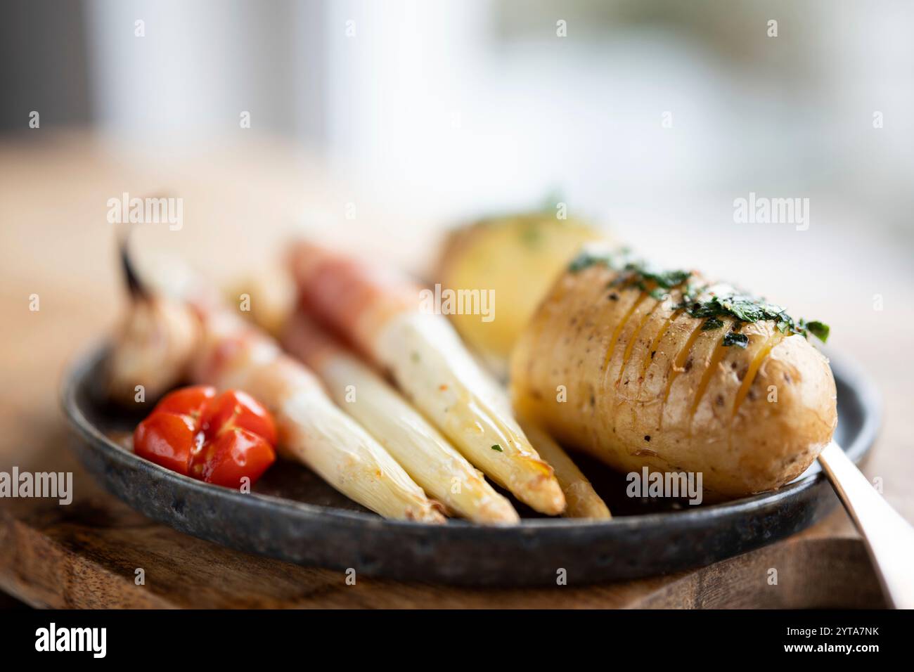 Asparagi bianchi fritti avvolti in pancetta con patate alle erbe e pomodori su piatto di ceramica nera e tavolo di legno. Profondità di campo ridotta. Foto Stock