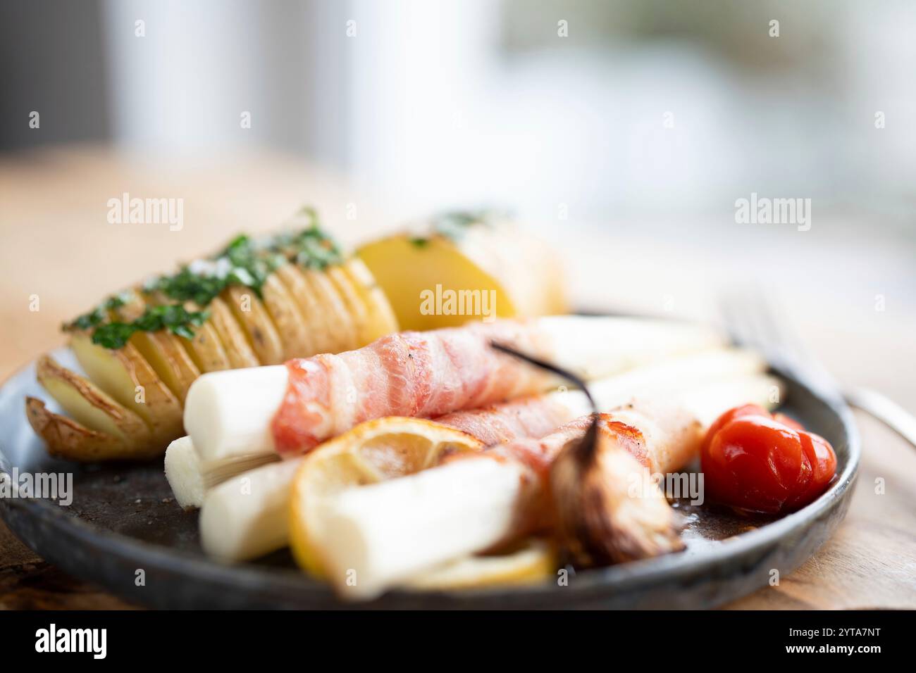 Asparagi bianchi fritti avvolti in pancetta con patate alle erbe e pomodori su piatto di ceramica nera e tavolo di legno. Profondità di campo ridotta. Foto Stock