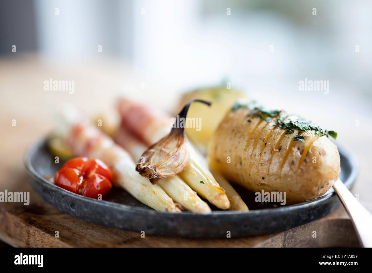 Asparagi bianchi fritti avvolti in pancetta con patate alle erbe e pomodori su un piatto di ceramica nera da vicino con poca profondità di campo. Foto Stock