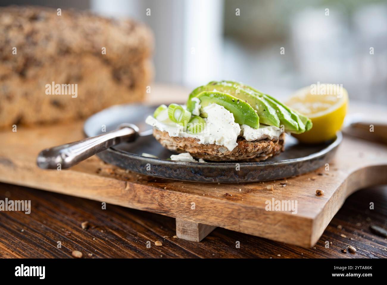 Fette di avocado fresco sul pane con formaggio spalmabile e limone. Cibo vegetariano su tagliere di legno. Primo piano di fronte alla finestra luminosa. Profondità di campo ridotta. Foto Stock