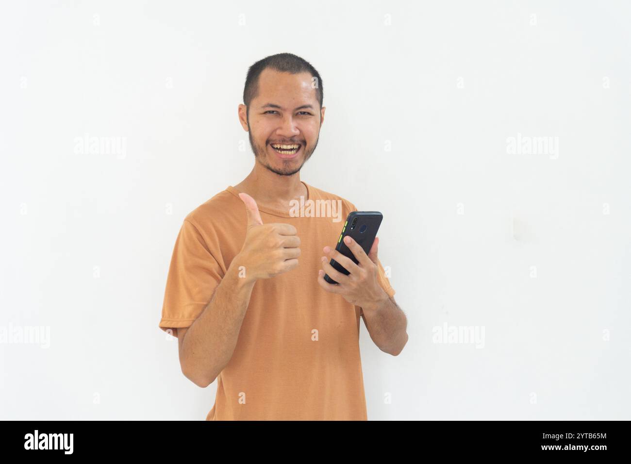 I giovani uomini che indossano una camicia arancione danno il pollice al sorriso del telefono cellulare su sfondo bianco isolato Foto Stock