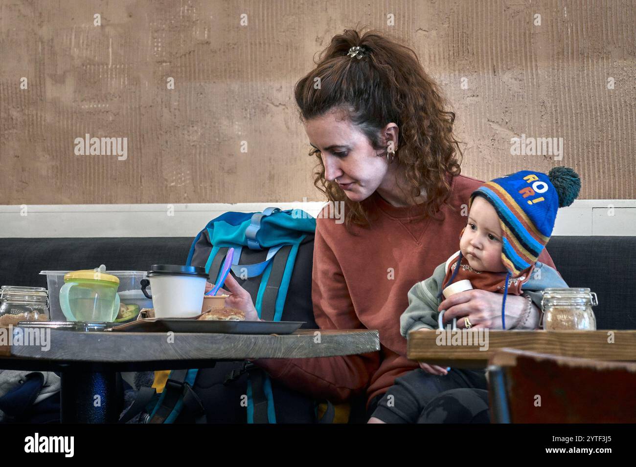 Madre multitasking con il bambino in braccio, che guarda lo smartphone mentre si nutre con il bambino in un ristorante Foto Stock