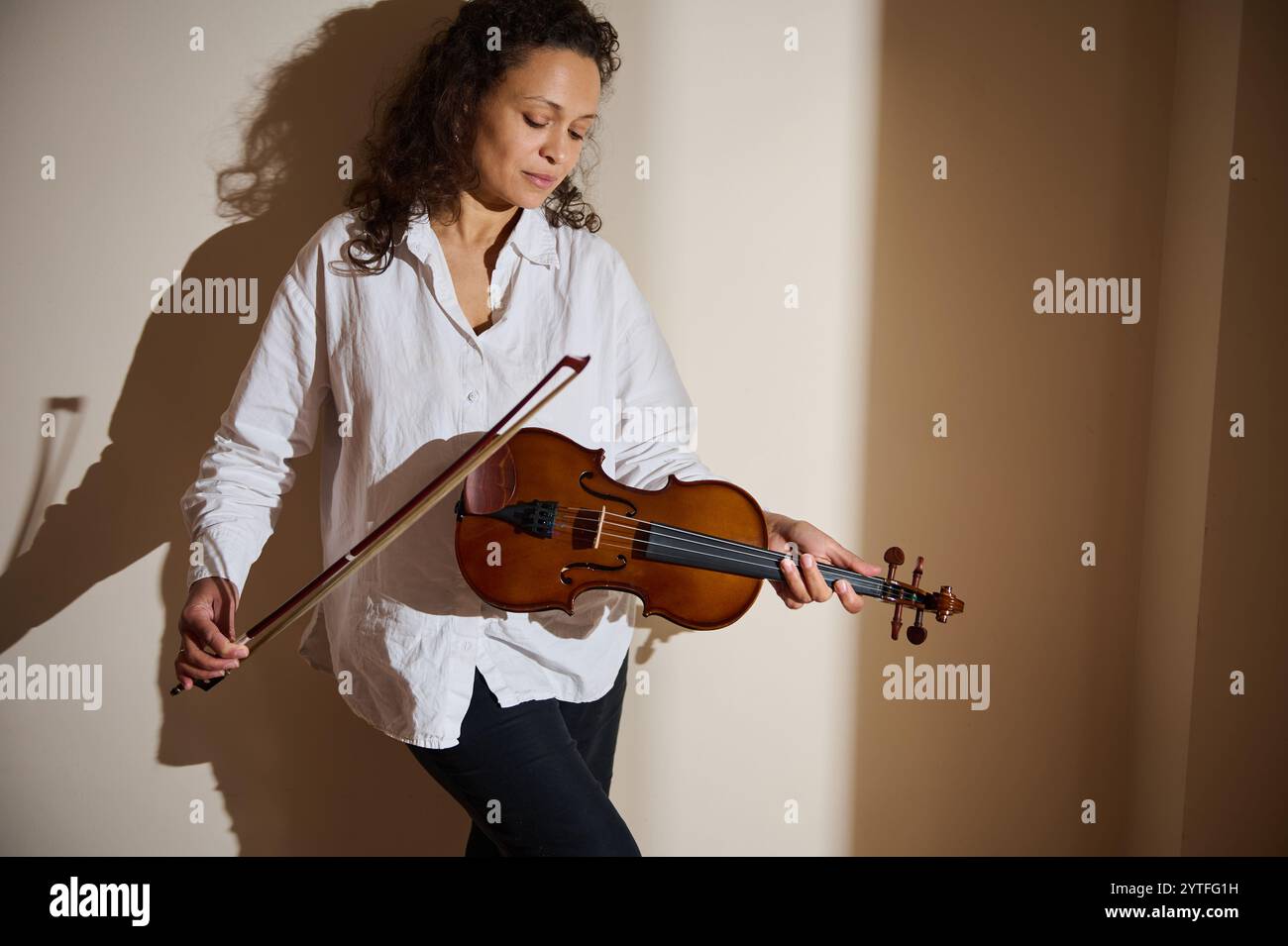 Una donna sta contro una parete illuminata, suona dolcemente un violino, immersa nella musica. Foto Stock