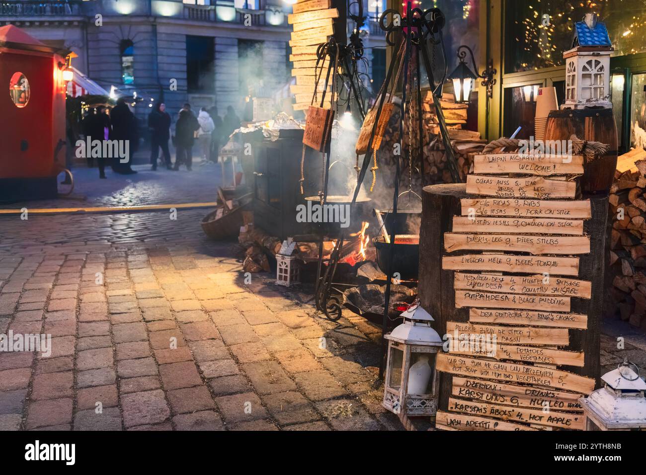 Cucina rustica al mercato di Natale di Piazza del Duomo con pentole di ferro appese su un fuoco aperto, tronchi di legno e lanterne su una strada acciottolata, crea Foto Stock