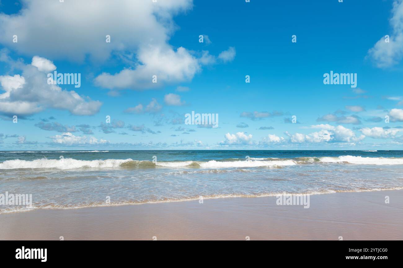 Paesaggio costiero con acque calme che toccano la sabbia, sotto un cielo blu segnato da nuvole bianche e grigie, creando un contrasto drammatico. Foto Stock