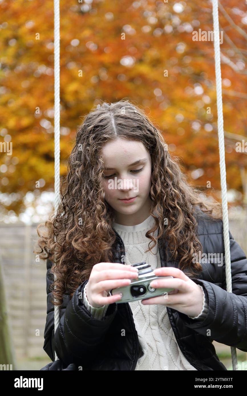 Adolescente che utilizza una fotocamera a pellicola da 35 mm analogica rétro in giardino seduto su un'altalena, controllando le impostazioni di apertura della velocità dell'otturatore della messa a fuoco Foto Stock