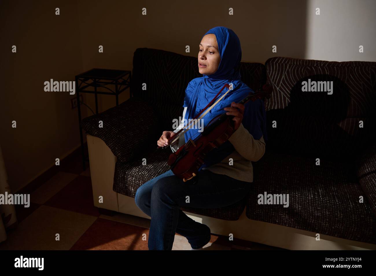 Una donna che indossa un hijab blu si siede su un divano, suona abilmente il violino, con luce e ombra che evidenziano la sua espressione focalizzata. Foto Stock