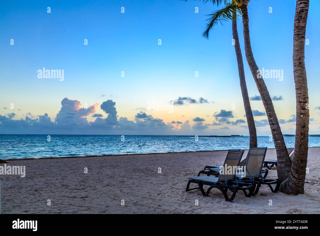 Una spiaggia con due sdraio e una palma. Il cielo è blu e il sole sta tramontando Foto Stock