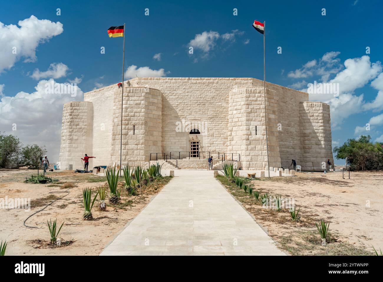 Il cimitero di guerra tedesco di El Alamein si erge come una fortezza sotto i cieli blu, con le bandiere della Germania e dell'Egitto sopra e la vegetazione che costeggia il percorso del deserto Foto Stock