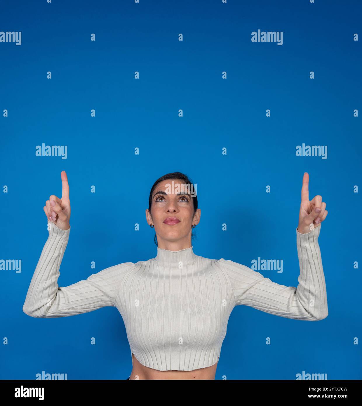Giovane donna che punta verso l'alto con entrambe le dita indice che guardano verso l'alto su uno sfondo blu Foto Stock