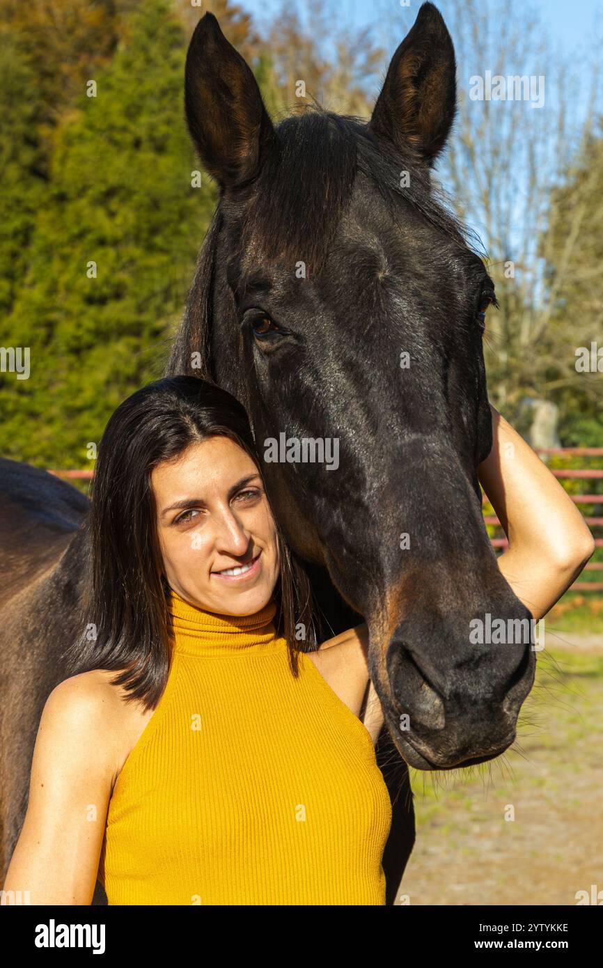 Primo piano di una donna in cima gialla sorridente mentre si trova accanto a un cavallo nero in un ranch all'aperto soleggiato, enfatizzando la compagnia e la campagna Foto Stock