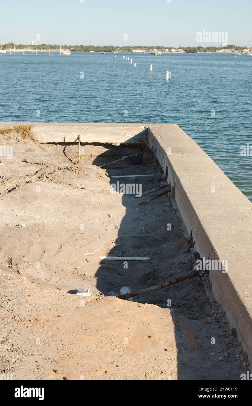 Vista d'angolo della diga danneggiata dalla tempesta erosa. Incrinature nei pannelli verticali della spalla e nel tappo. Calme acque blu della baia nella parte posteriore in una giornata di sole. Bianco e. Foto Stock