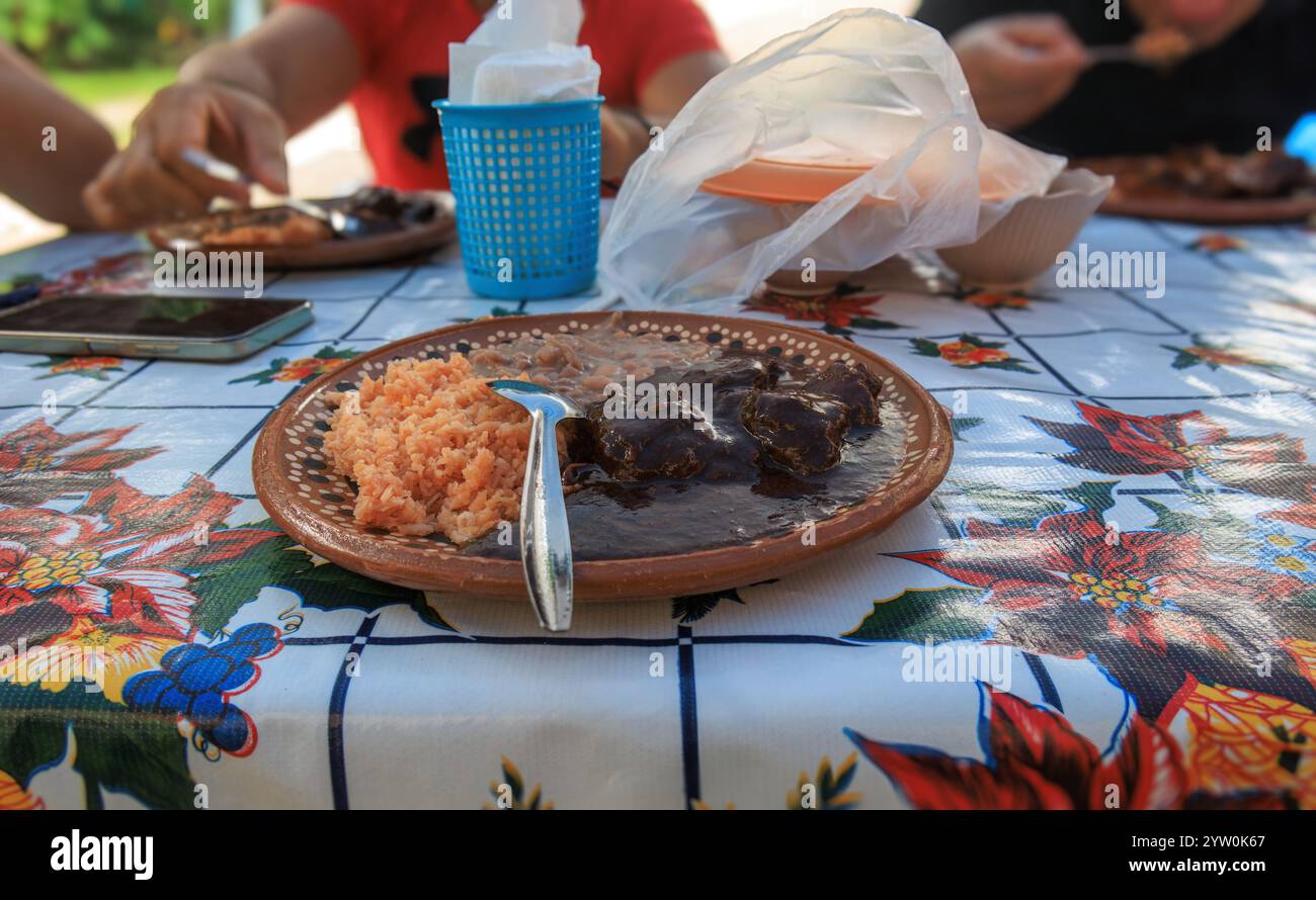 Un gruppo di persone si riunisce intorno a un tavolo colorato gustando un pasto tradizionale. L'attenzione è concentrata su un piatto caratterizzato da una ricca salsa scura che accompagna un mou Foto Stock