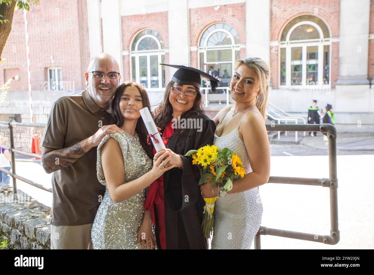 Famiglia che celebra la laurea di una donna all'esterno di un edificio in mattoni con grandi finestre e girasoli in mano, Bournemouth, Dorset, Regno Unito Foto Stock