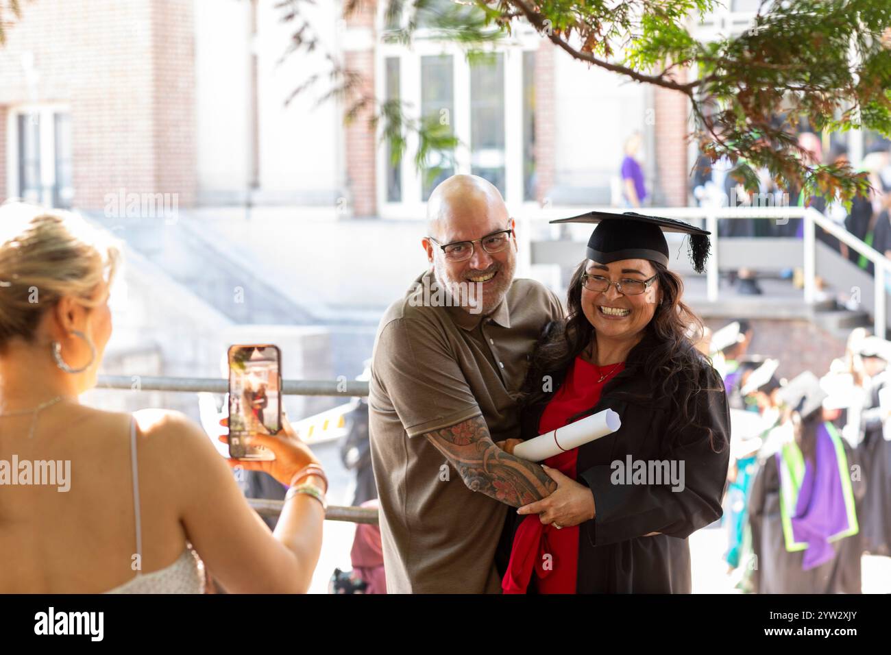 Festa del giorno della laurea con un uomo orgoglioso che abbraccia una donna in berretto e abito mentre un'altra persona scatta la propria foto, Bournemouth, Dorset UK Foto Stock