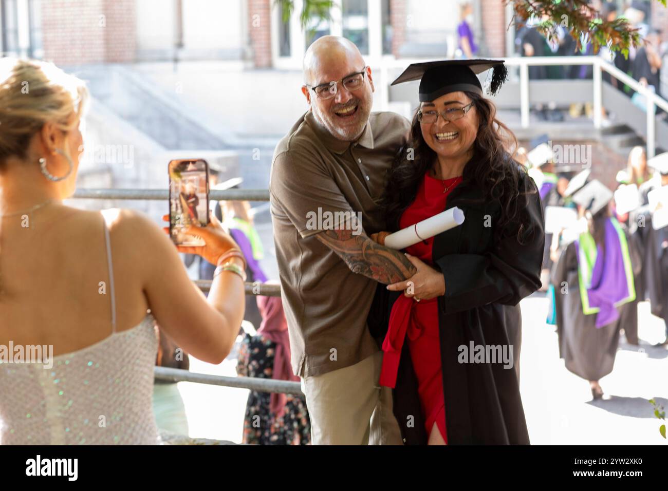 Una laureata sorridente in berretto e abito abbraccia un uomo mentre posa per una foto il giorno della sua laurea, Bournemouth, Dorset UK Foto Stock