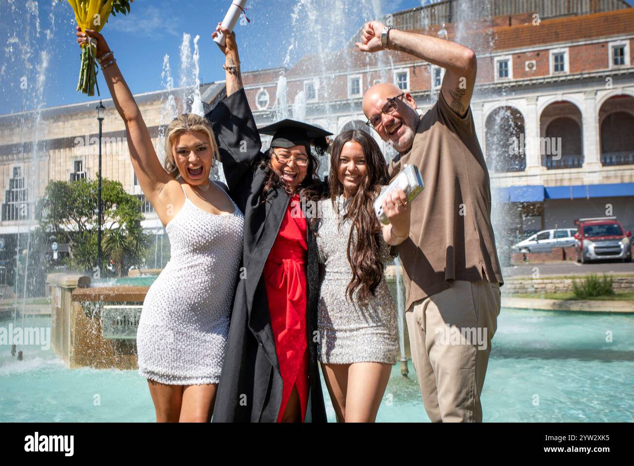Gruppo di persone gioiose che celebrano la laurea con una giovane donna in un berretto e abito, alzando il diploma vicino a una fontana in una giornata di sole, Bournemouth, Dorset UK Foto Stock