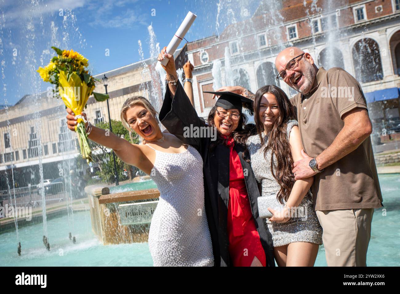 Gruppo di persone felici che festeggiano una laurea con un bouquet e un diploma vicino a una fontana in una giornata di sole, Bournemouth, Dorset, Regno Unito Foto Stock