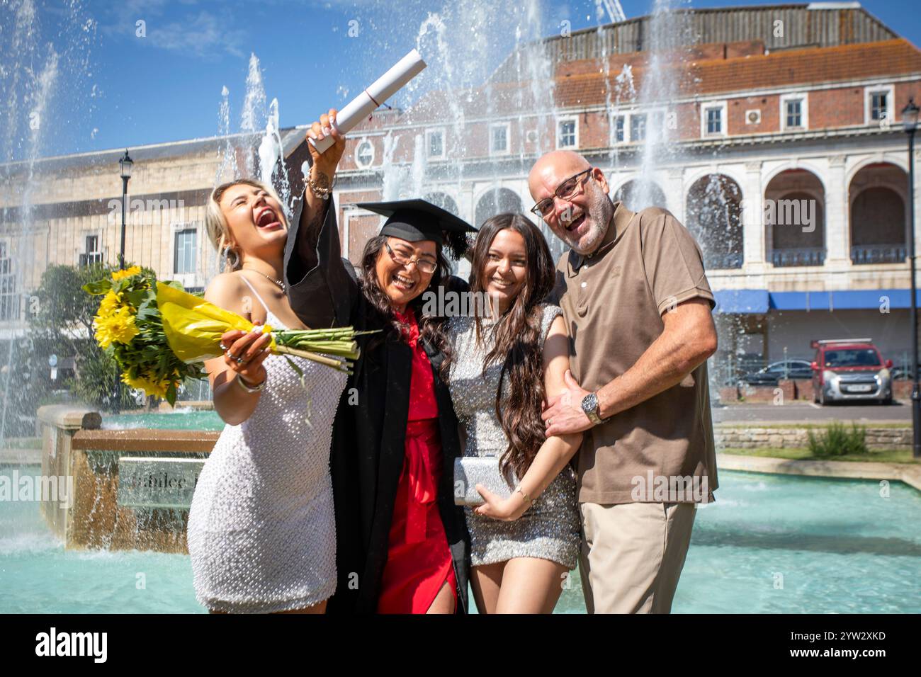 Celebrazione della laurea con un felice laureato che tiene un diploma e fiori, posando con amici e familiari vicino a una fontana in una giornata di sole, Bournemouth, Dorset, Regno Unito Foto Stock