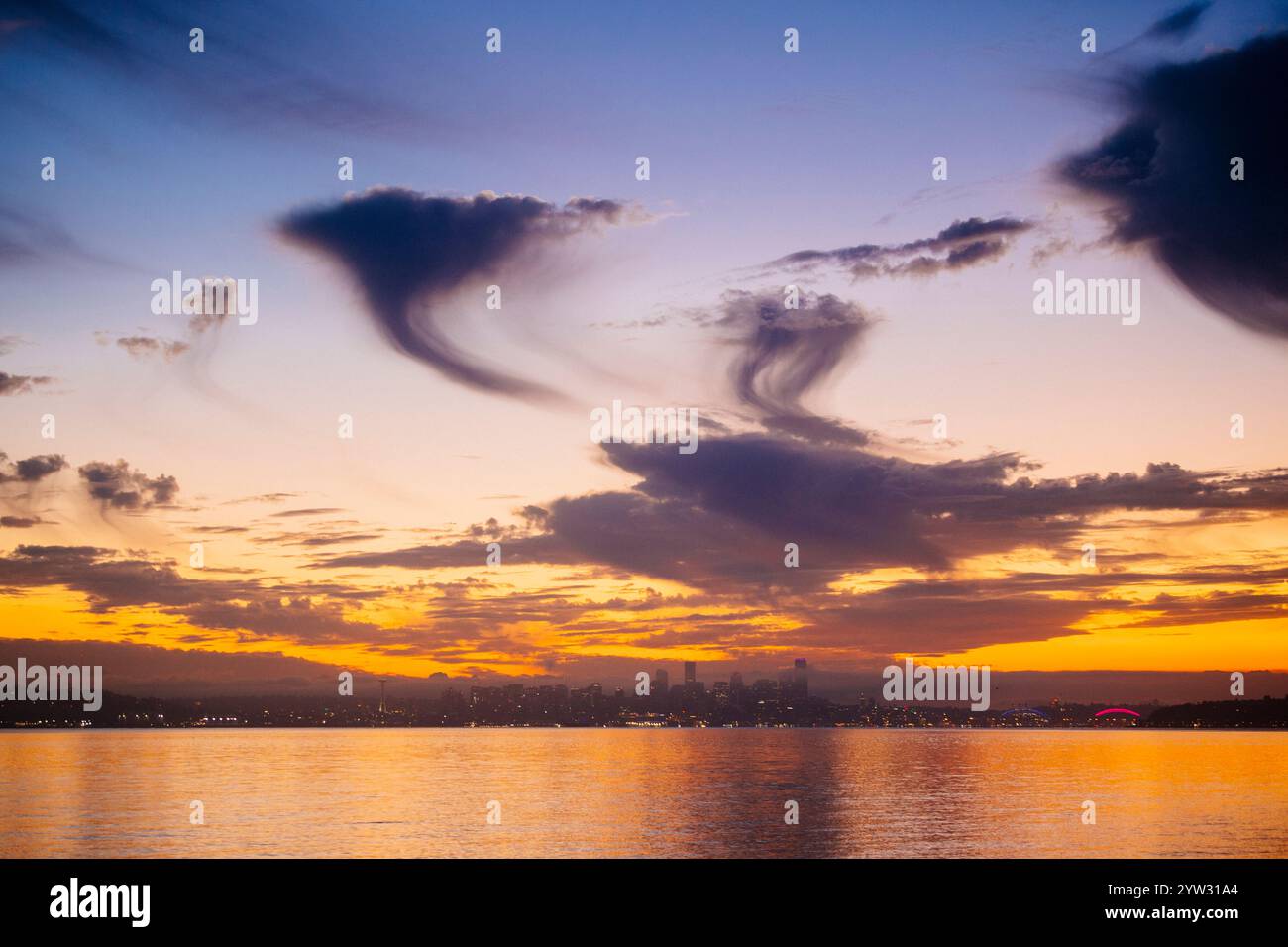 Cielo al tramonto con spettacolari nuvole su un calmo corpo d'acqua con una silhouette dello skyline della città in lontananza. Foto Stock