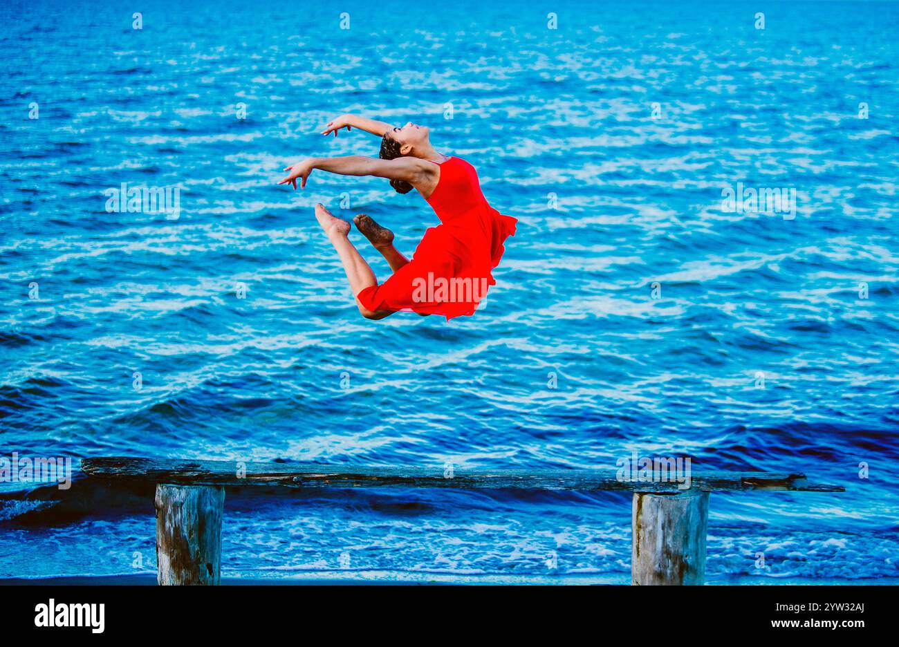 Graziosa ballerina in abito rosso che esegue un salto sull'acqua blu con una struttura in legno in primo piano, Stati Uniti Foto Stock