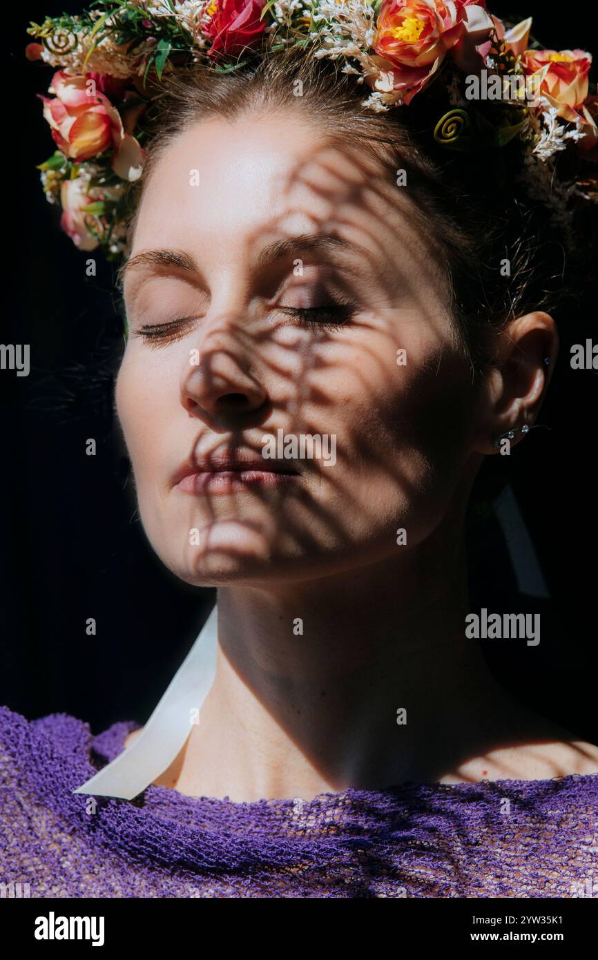 Una donna serena con una corona floreale e gli occhi chiusi, bagnata dalla luce del sole con motivi d'ombra sul viso, USA Foto Stock