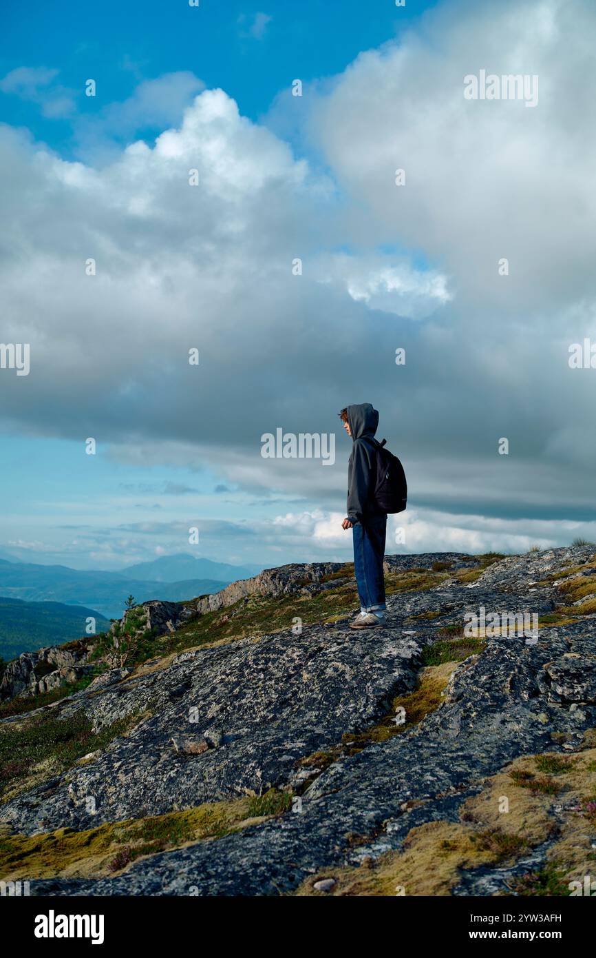 Un adolescente in una felpa con cappuccio e zaino in piedi in cima a una collina rocciosa, che guarda il cielo nuvoloso e le montagne lontane, Romsdalfjorden, Norvegia Foto Stock
