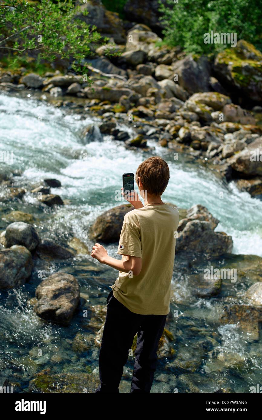Adolescente in abiti casual che utilizza uno smartphone per catturare un momento da un torrente roccioso di montagna, Trollstiege, Trollvegen, Norvegia Foto Stock Adolescente in abiti casual che utilizza uno smartphone per catturare un momento da un torrente roccioso di montagna, Trollstiege, Trollvegen, Norvegia Foto Stock