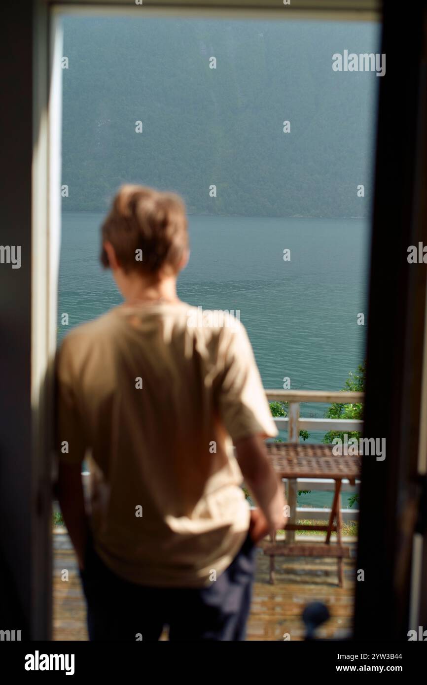 Adolescente in piedi ad una porta aperta che guarda un tranquillo lago e montagne, Storfjorden, Norvegia Foto Stock