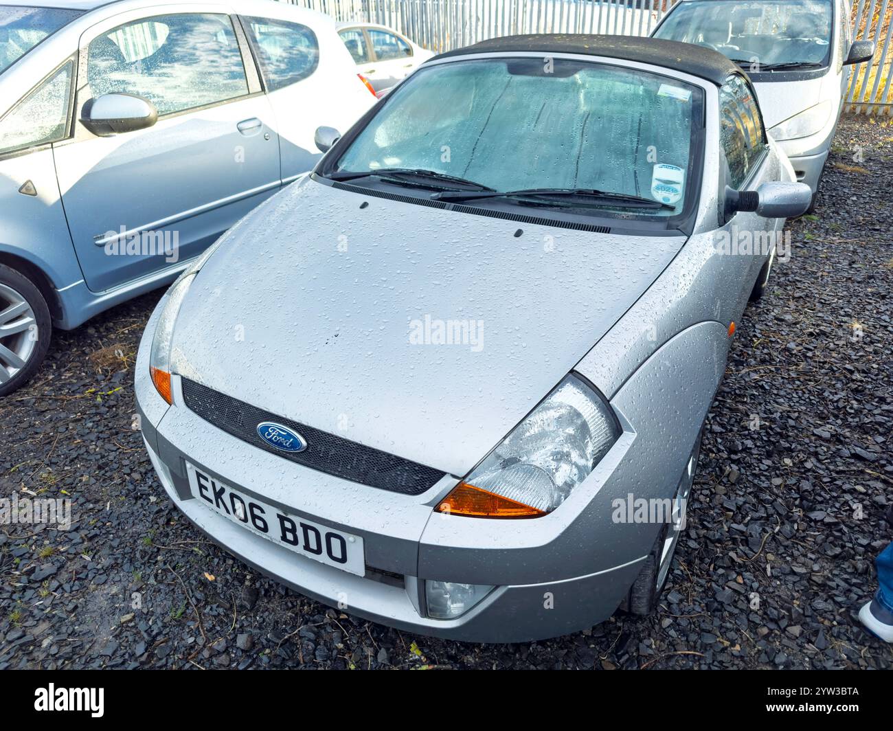 Una Ford KA convertibile compatta color argento parcheggiata su un terreno ghiaioso tra la vista anteriore di altri veicoli in una giornata nuvolosa Foto Stock