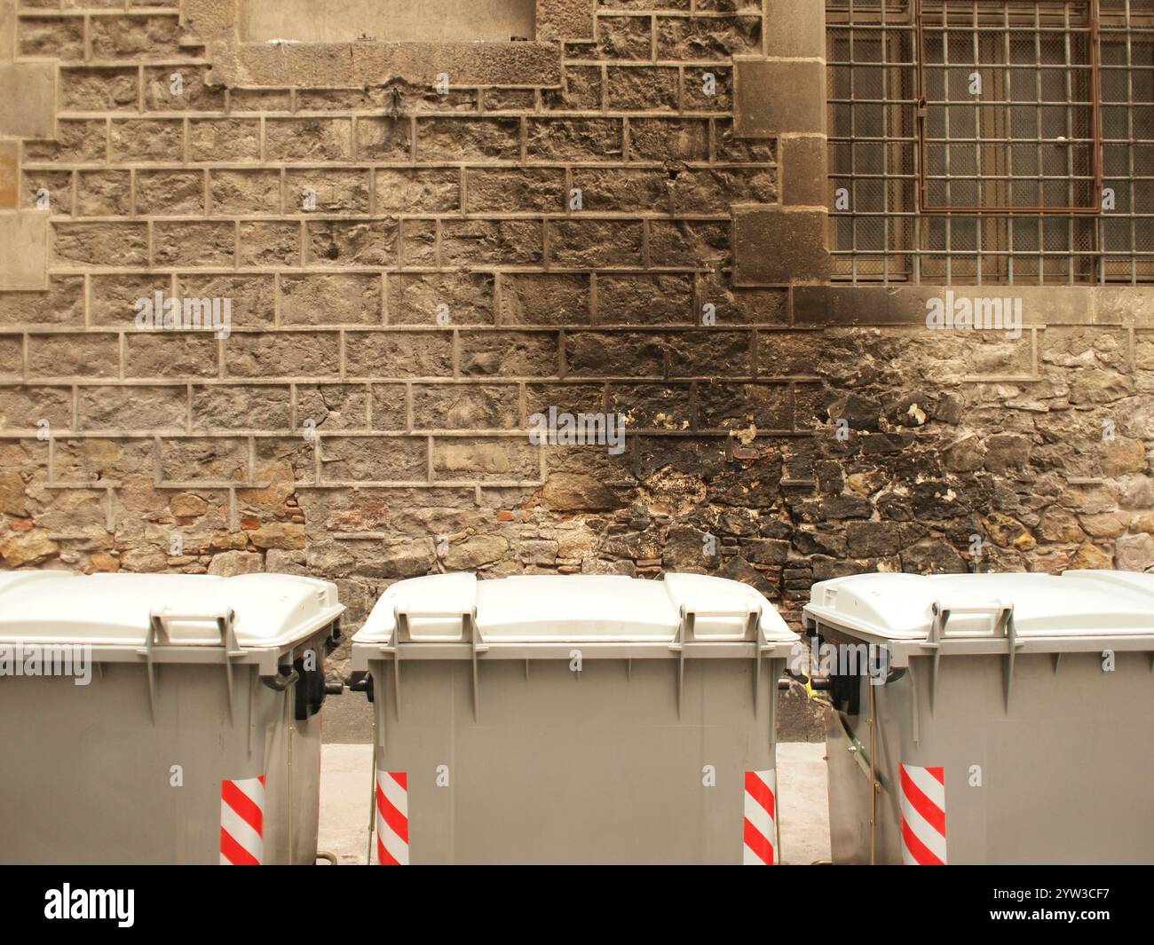 Tre bidoni grigi della spazzatura allineati contro un vecchio muro di pietra con una griglia di metallo e tracce brucianti sopra di loro, Barcellona, Spagna Foto Stock