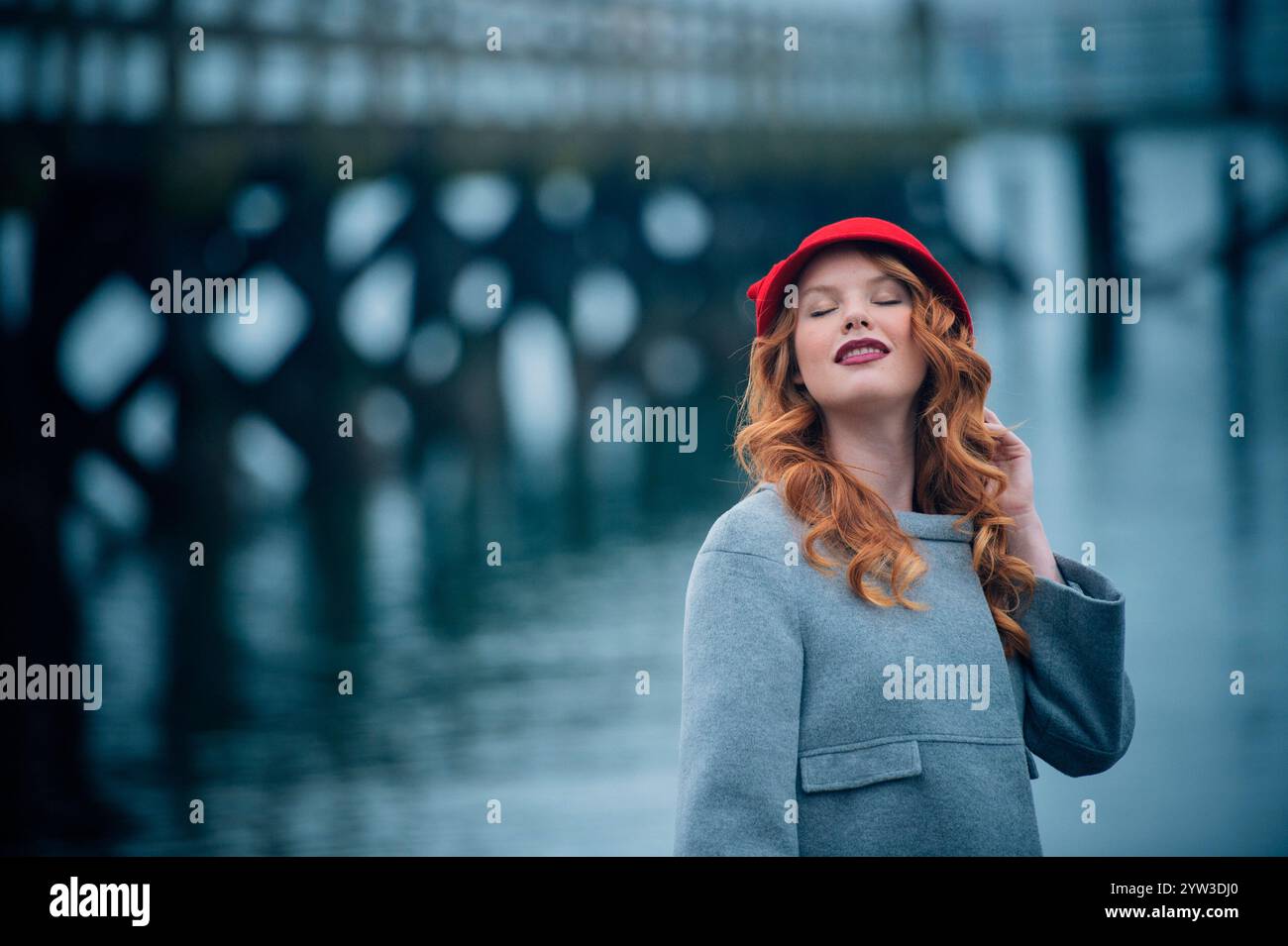 Una donna rossa sorridente con un cappello rosso e un cappotto grigio in piedi di fronte a un ponte sull'acqua. Foto Stock