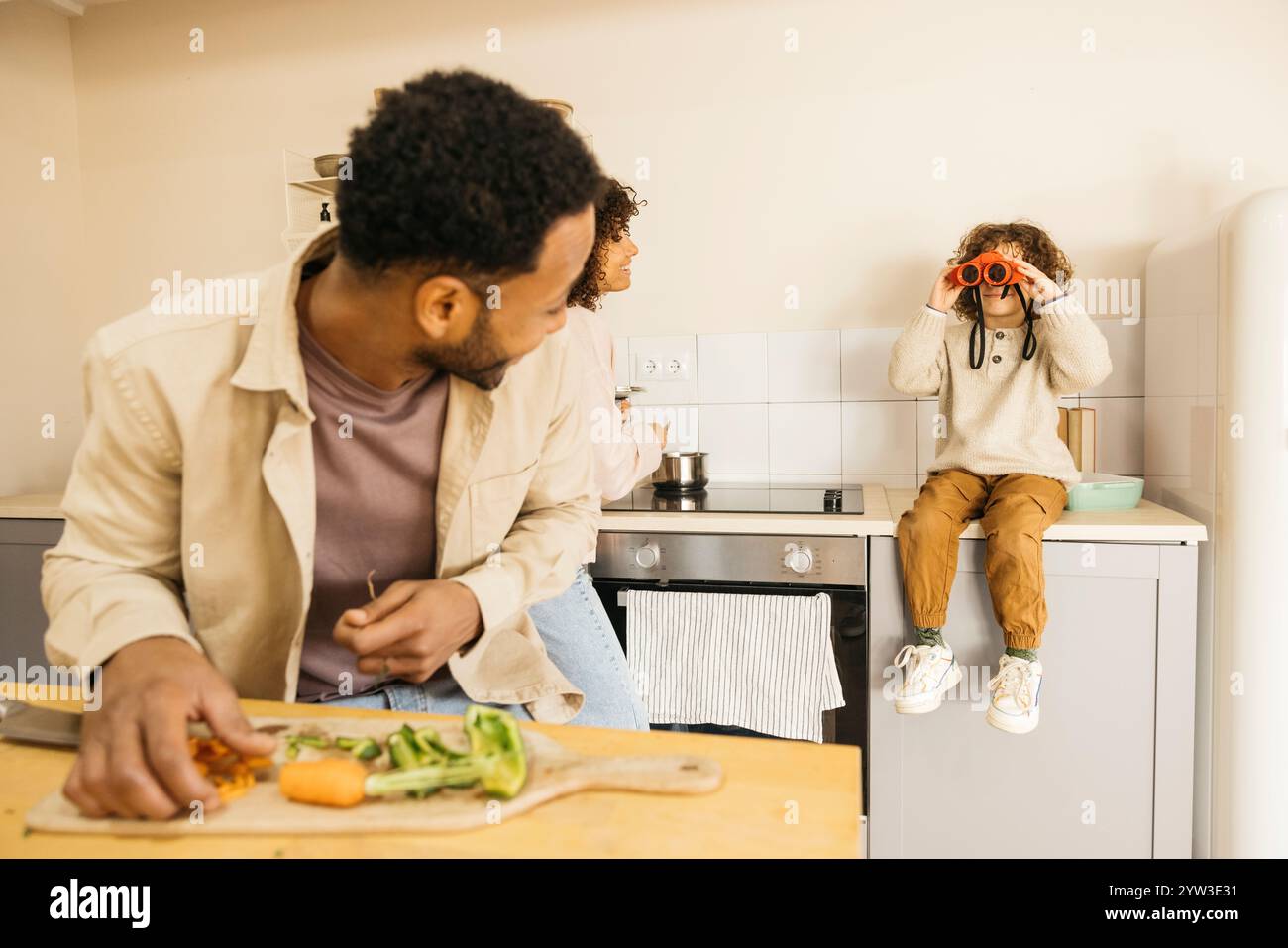 Una famiglia condivide momenti di gioia in cucina. Un bambino gioca con il binocolo mentre un adulto prepara le verdure, creando un'atmosfera calda e giocosa. Foto Stock