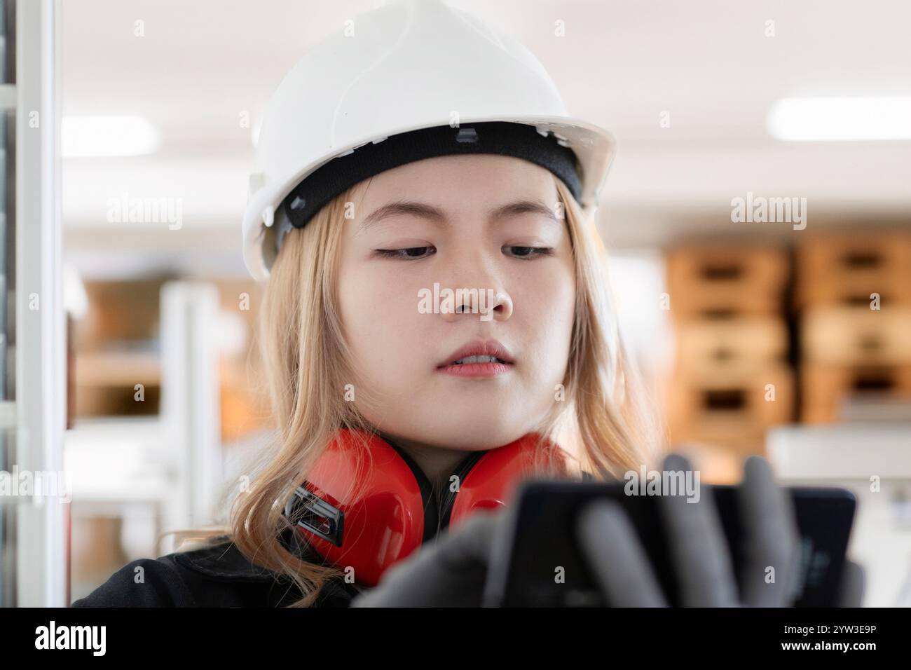 Una giovane ingegnere che indossa un casco di sicurezza bianco e una protezione per le orecchie arancione esamina un dispositivo in mano in ambienti chiusi, Germania Foto Stock