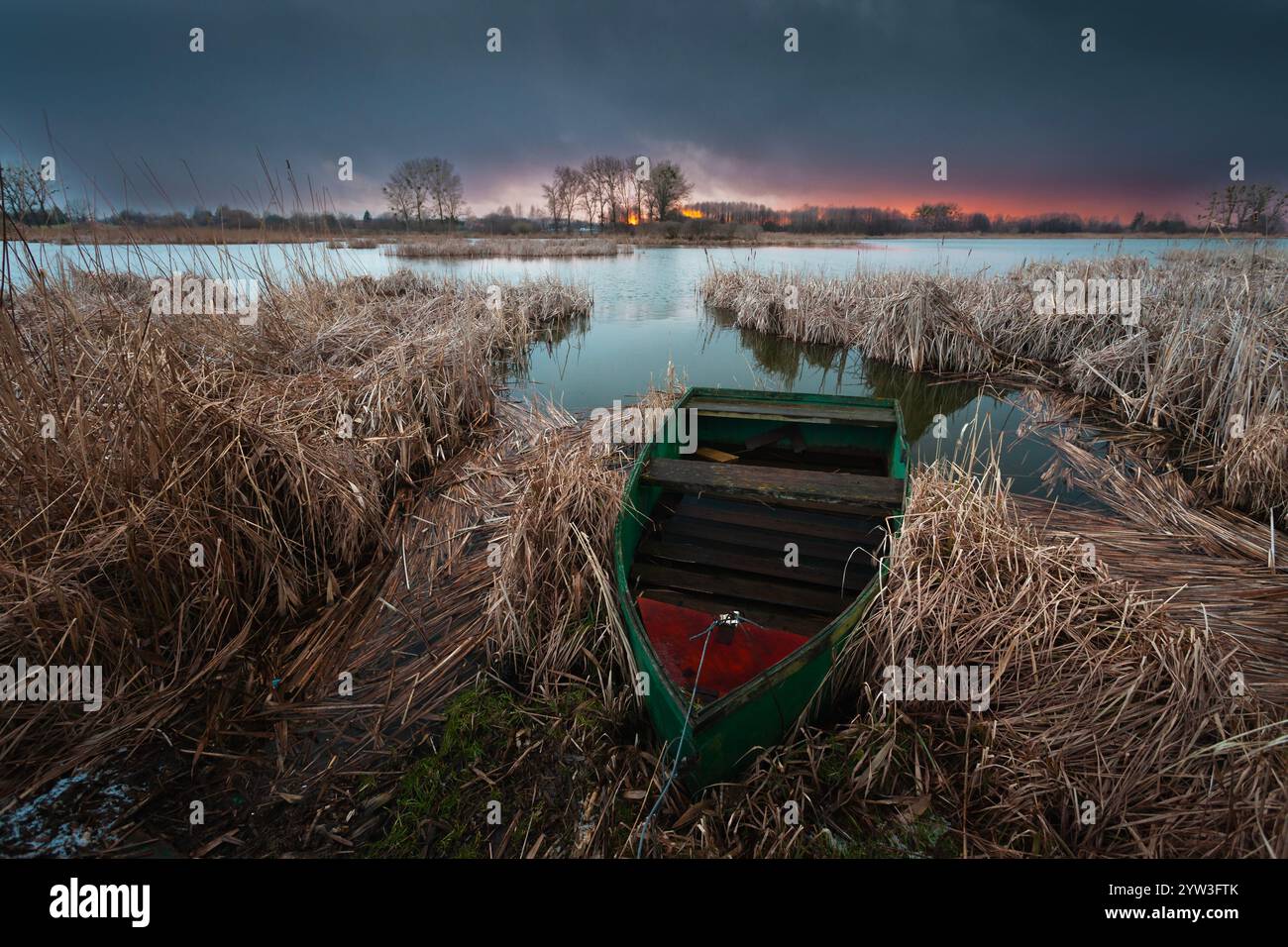 Una vista di una vecchia barca da pesca ormeggiata nelle canne asciutte sulla riva di un lago durante un tramonto nuvoloso. Foto Stock