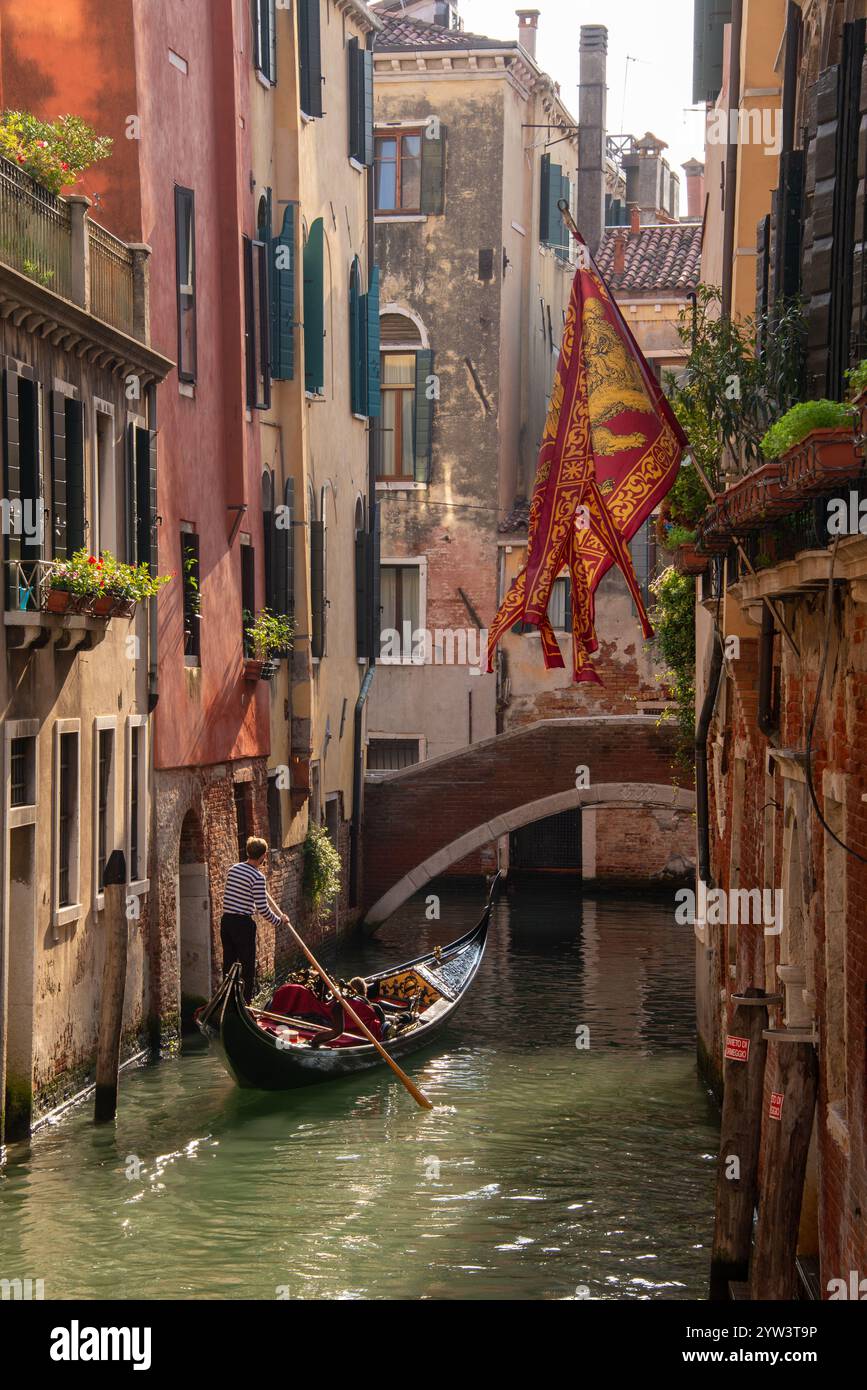 Venezia classica: Una bandiera con il Leone di San Marco, il santo patrono di Venezia che sorvola uno dei canali più piccoli e una gondola trainata da un gondoliere in uno dei tanti canali più piccoli, i cosiddetti "rios" che attraversano il centro storico. A causa della costante umidità, la maggior parte dei vecchi edifici hanno un aspetto piuttosto romantico, a volte morboso. Il centro storico di Venezia è patrimonio dell'umanità dell'UNESCO. Foto Stock