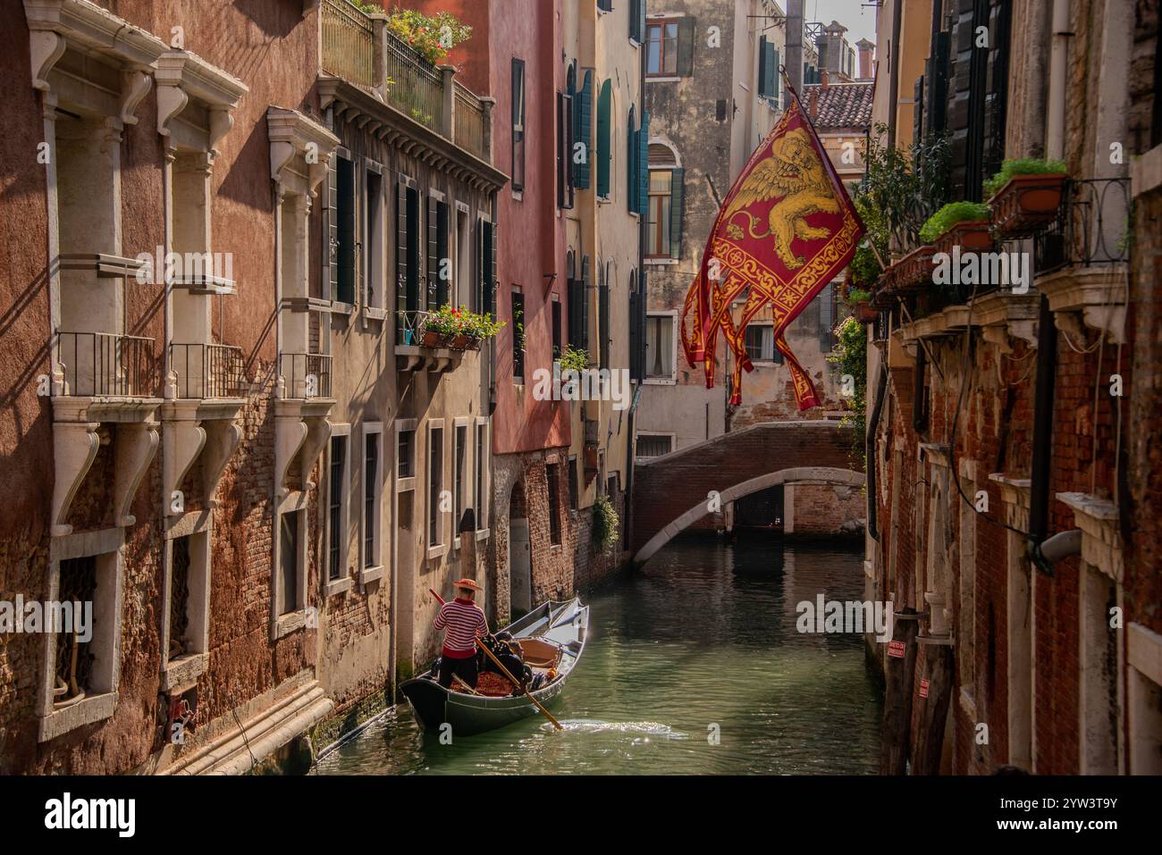 Venezia classica: Una bandiera con il Leone di San Marco, il santo patrono di Venezia che sorvola uno dei canali più piccoli e una gondola trainata da un gondoliere in uno dei tanti canali più piccoli, i cosiddetti "rios" che attraversano il centro storico. A causa della costante umidità, la maggior parte dei vecchi edifici hanno un aspetto piuttosto romantico, a volte morboso. Il centro storico di Venezia è patrimonio dell'umanità dell'UNESCO. Foto Stock