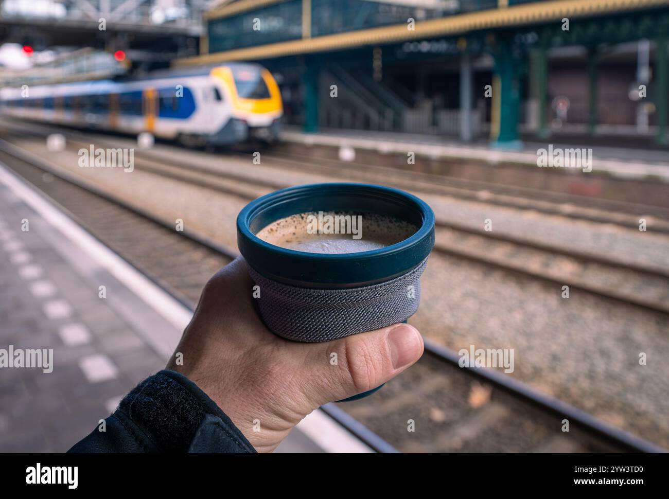 Gusta un caffè alla stazione ferroviaria olandese, tenendo in mano una tazza di cappuccino riutilizzabile e pieghevole in plastica mentre aspetti il treno Foto Stock