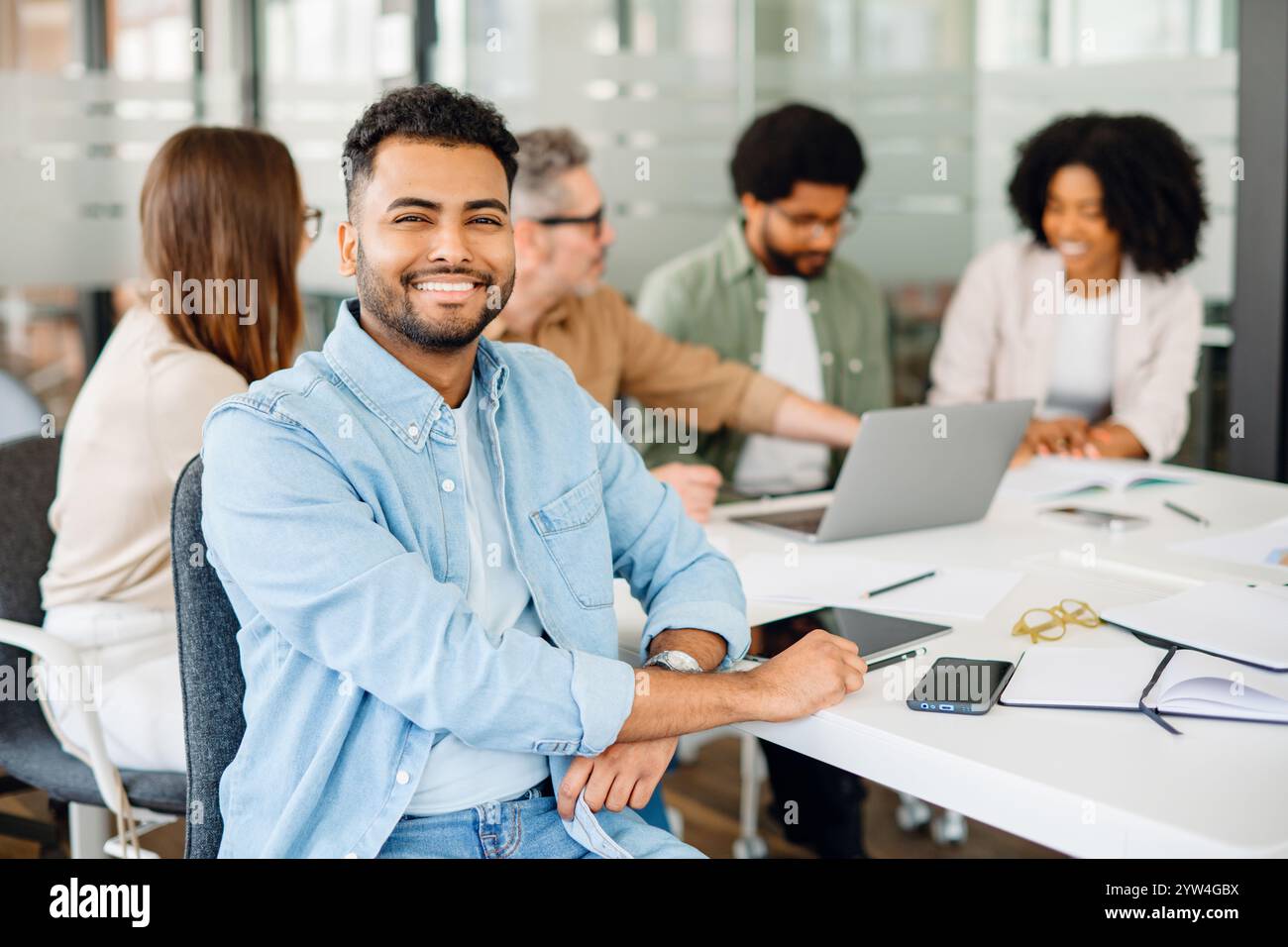 Un giovane con una camicia blu sorride con fiducia alla telecamera, con i suoi colleghi impegnati in una discussione sullo sfondo, mostrando un team armonioso in un ambiente produttivo Foto Stock