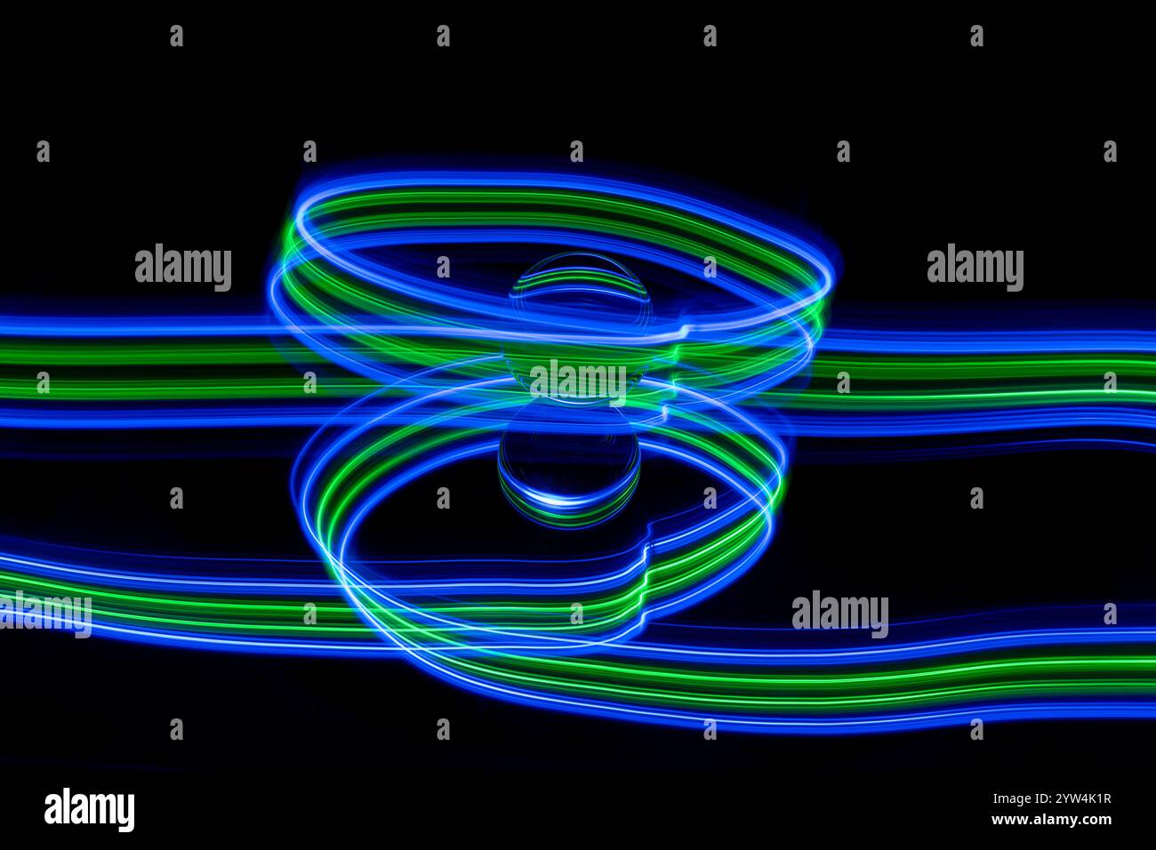 Tracce di luce blu e verde astratte create dalla pittura leggera intorno a una sfera di vetro statica posizionata su uno specchio su uno sfondo nero semplice Foto Stock