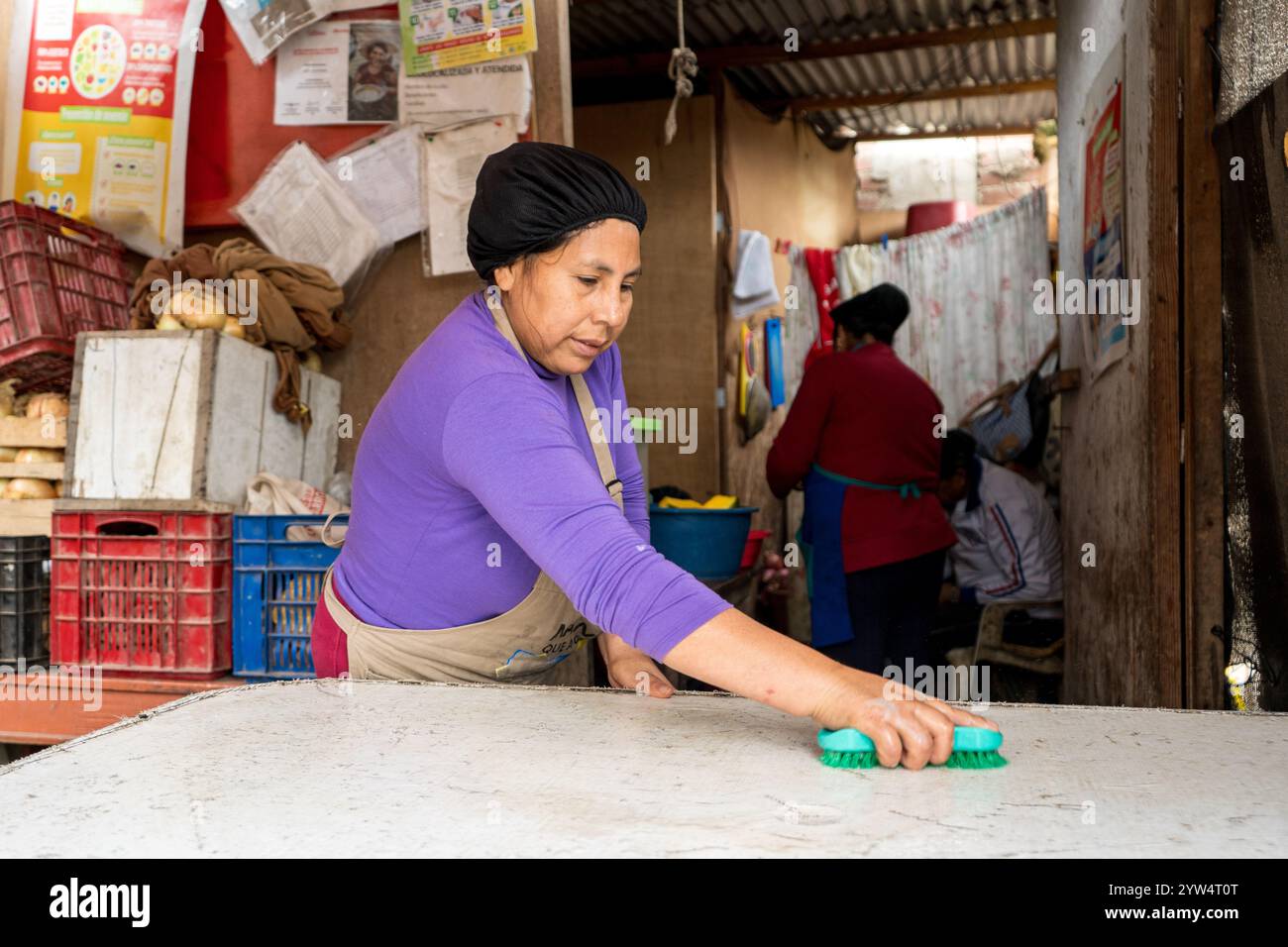 Lurin, Perù - 6 settembre 2024: Young Woman in a Purple Shirt and Kitchen Apron Cleans a White Table in the Modest Community Kitchen Foto Stock