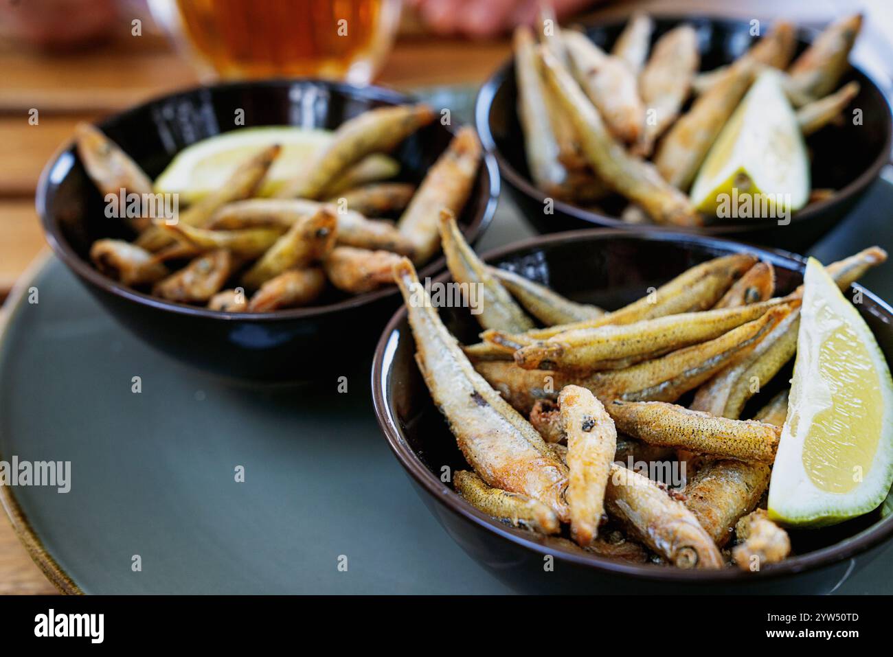 Piccolo pesce fritto croccante servito con spicchi di limone in ciotole scure su un tavolo di legno, accompagnato da un bicchiere di birra sullo sfondo. Un informale, appetizi Foto Stock