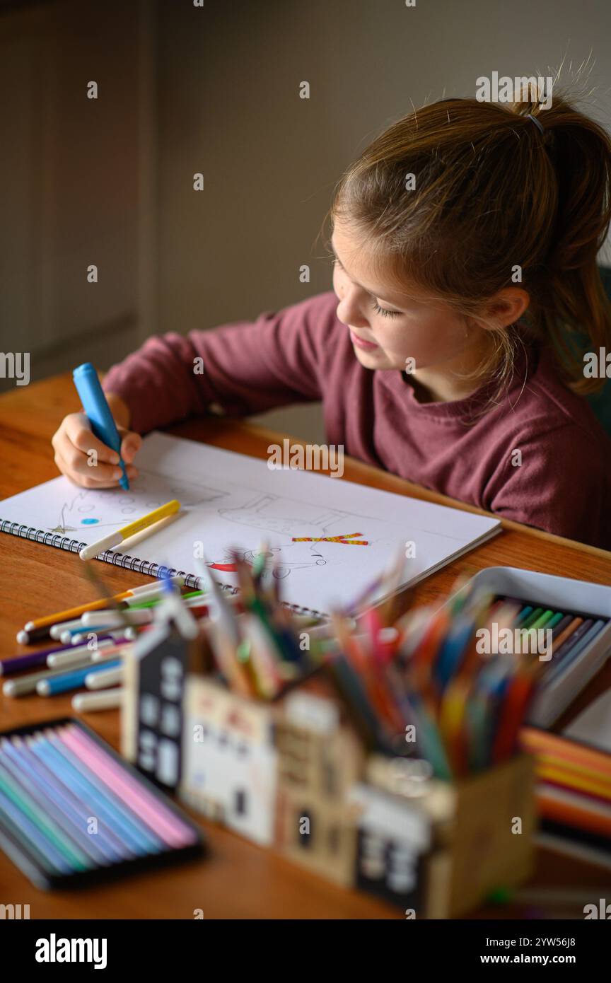 la ragazza è seduta sul tavolo che sta studiando pittura e lettura con molti pennarelli e matite intorno Foto Stock
