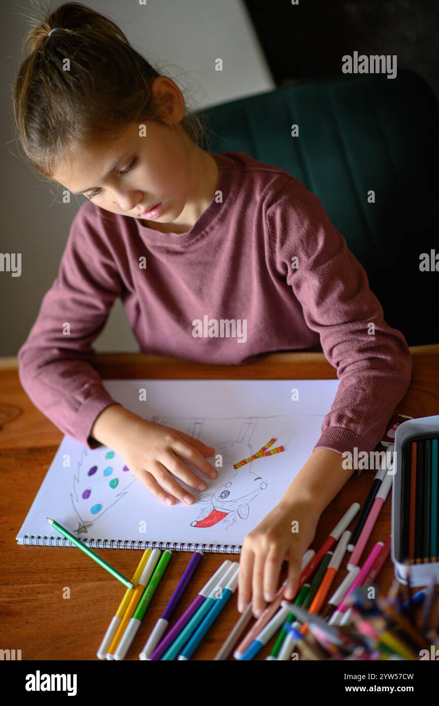 la ragazza è seduta sul tavolo che sta studiando pittura e lettura con molti pennarelli e matite intorno Foto Stock