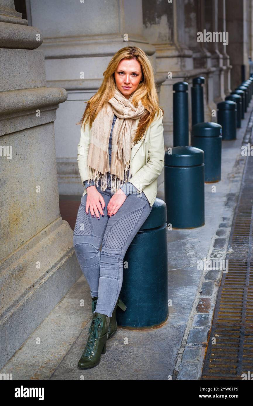 Una donna con lunghi capelli biondi e una sciarpa beige si siede casualmente contro un grande pilastro di pietra. È vestita con una giacca di colore chiaro, pantaloni grigi e. Foto Stock