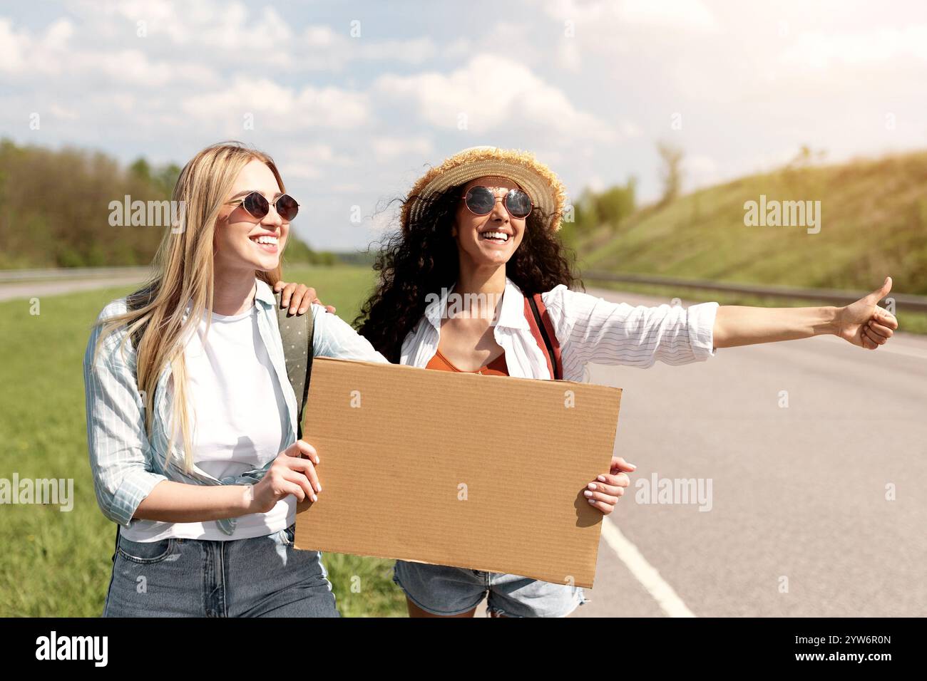 Due amiche che si divertono a cavalcare liberamente, tenendo un cartello di cartone vuoto con il mockup, fermando l'auto su strada, spazio libero Foto Stock