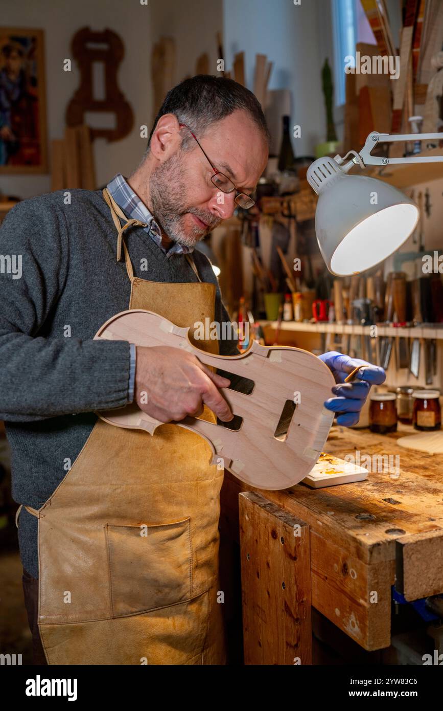 Cremona, Italia - 9 dicembre 2024 - Luca tenore maestro liutaio che lavora su un violino nella sua bottega, applicando la vernice con un pennello Foto Stock