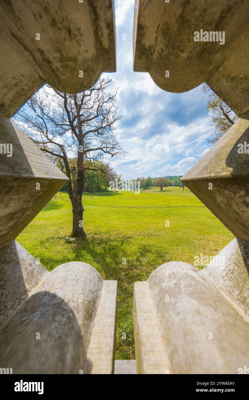 Splendido paesaggio di un grande parco verde con radure verdi, alberi al mattino nuvoloso e soleggiato visti attraverso un buco decorativo in cemento recinzione di un piccolo ponte Foto Stock