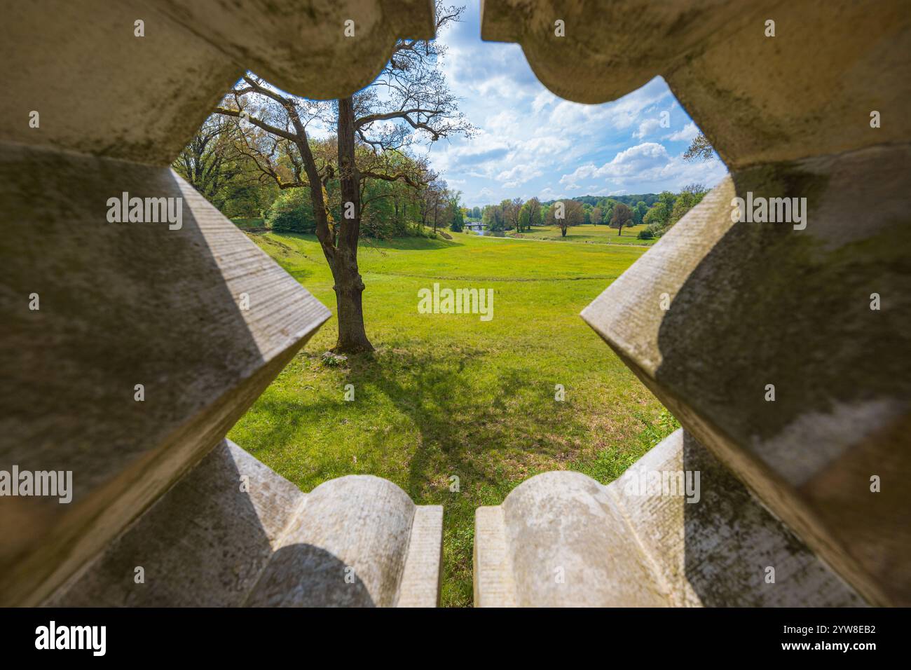 Splendido paesaggio di un grande parco verde con radure verdi, alberi al mattino nuvoloso e soleggiato visti attraverso un buco decorativo in cemento recinzione di un piccolo ponte Foto Stock