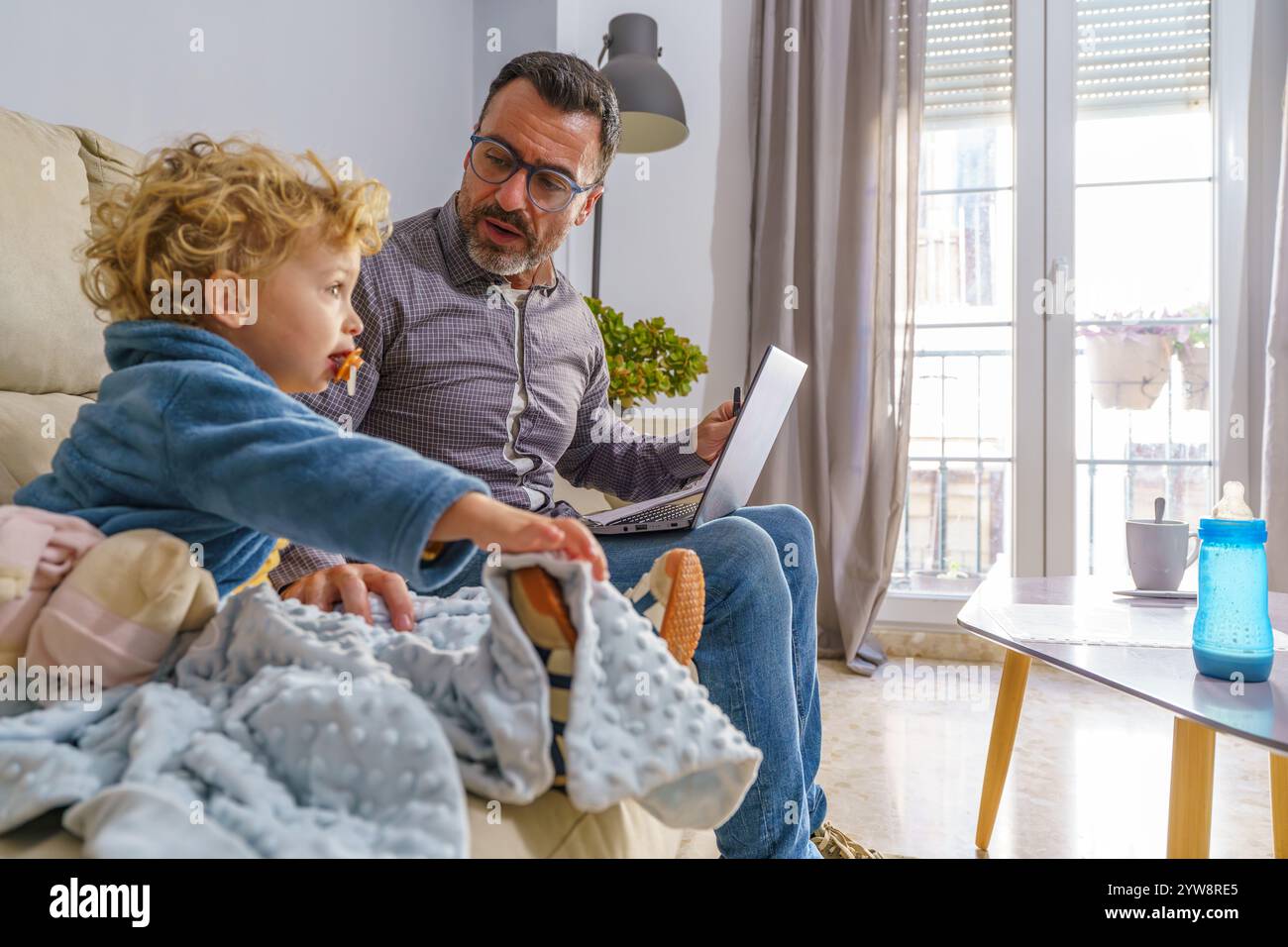 Padre che lavora in multitasking con il lavoro e la famiglia, utilizzando un notebook mentre si prende cura del bambino in un comodo soggiorno Foto Stock